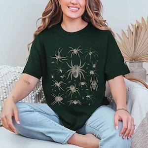 May include: A dark forest green short-sleeved t-shirt featuring a central graphic print of various spider species in a light beige colour. The design displays multiple arachnids of different sizes and shapes arranged in a grid-like pattern on the chest.