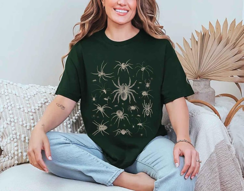 May include: A dark forest green short-sleeved t-shirt featuring a central graphic print of various spider species in a light beige colour. The design displays multiple arachnids of different sizes and shapes arranged in a grid-like pattern on the chest.