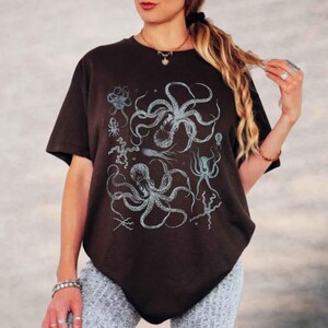 May include: A dark brown short-sleeved t-shirt featuring a detailed graphic print of various sea creatures, including two large octopuses, a squid, and smaller marine life, all rendered in a light blue and white illustrative style on the front.
