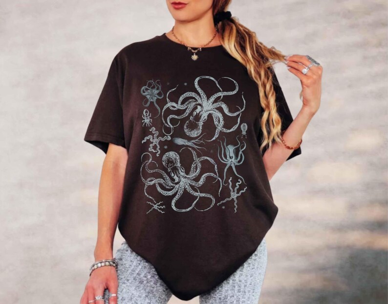 May include: A dark brown short-sleeved t-shirt featuring a detailed graphic print of various sea creatures, including two large octopuses, a squid, and smaller marine life, all rendered in a light blue and white illustrative style on the front.