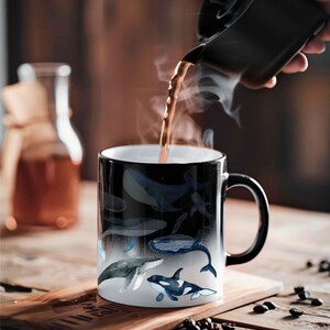 May include: A black ceramic mug with a white interior, featuring a whale design. The mug is being filled with a dark liquid, creating steam. The mug is on a wooden surface.
