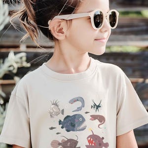 May include: A beige short-sleeved t-shirt featuring a graphic print of various deep-sea creatures, including an anglerfish, a viperfish, and other marine life in shades of blue, grey, and red, worn with light-coloured sunglasses.