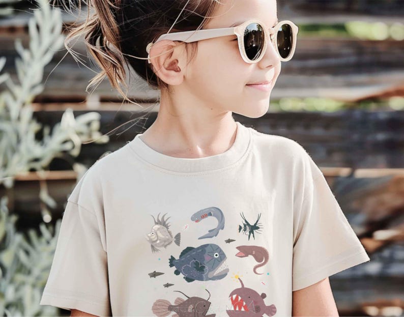 May include: A beige short-sleeved t-shirt featuring a graphic print of various deep-sea creatures, including an anglerfish, a viperfish, and other marine life in shades of blue, grey, and red, worn with light-coloured sunglasses.