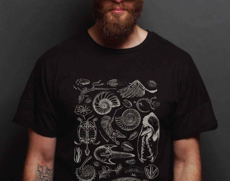 May include: A black short-sleeved t-shirt featuring a white graphic print of various fossils, including ammonites, a turtle skeleton, a bird skull, and other prehistoric biological specimens arranged in a detailed, scientific illustration style.