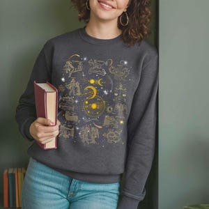 May include: A dark gray sweatshirt with a gold zodiac constellation print. The print features all 12 zodiac signs, the sun, moon, and stars.