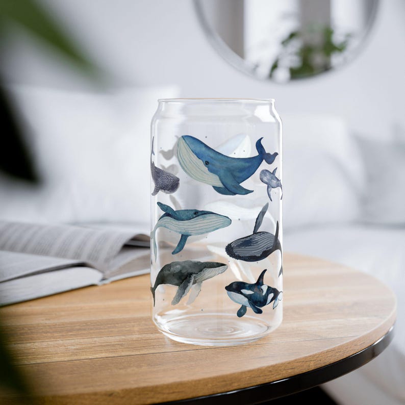 May include: A clear glass can-shaped drinking glass decorated with watercolour illustrations of various whales in shades of blue and grey. The whales are swimming around the glass. The glass is on a wooden table.