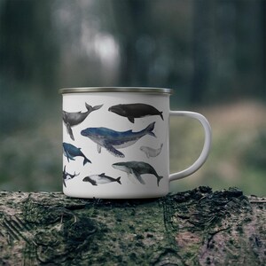 Peut inclure: Mug de camping en &eacute;mail blanc avec une illustration color&eacute;e de diff&eacute;rentes esp&egrave;ces de baleines. Le mug a une poign&eacute;e noire et un bord argent&eacute;.