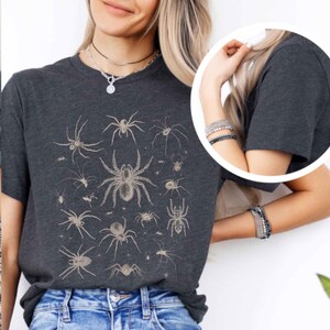 May include: A dark charcoal grey short-sleeve t-shirt featuring a detailed graphic print of various spider species in a light beige colour. The design displays multiple spiders of different sizes and shapes arranged across the front of the shirt.