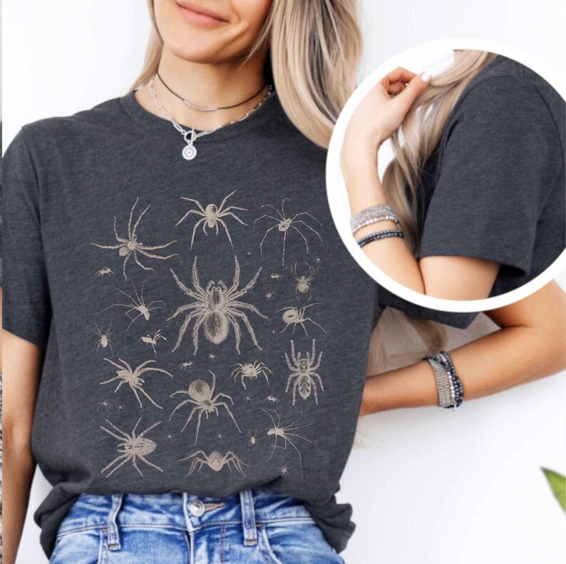 May include: A dark charcoal grey short-sleeve t-shirt featuring a detailed graphic print of various spider species in a light beige colour. The design displays multiple spiders of different sizes and shapes arranged across the front of the shirt.