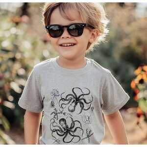 May include: A light gray short-sleeved t-shirt featuring a detailed black ink-style graphic print of multiple octopuses and various sea creatures arranged across the front of the garment, with a crew neckline and a standard fit.