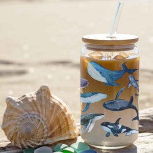 May include: A glass tumbler with a bamboo lid and straw, adorned with watercolour illustrations of whales. The tumbler contains a light brown liquid. A seashell rests beside it on a sandy surface.