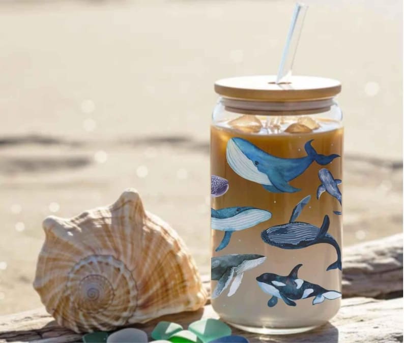 May include: A glass tumbler with a bamboo lid and straw, adorned with watercolour illustrations of whales. The tumbler contains a light brown liquid. A seashell rests beside it on a sandy surface.