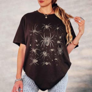 May include: A black short-sleeved t-shirt featuring a graphic print of various spiders in a light beige colour. The design displays multiple spider species of different sizes arranged across the front of the garment, which has a crew neckline.