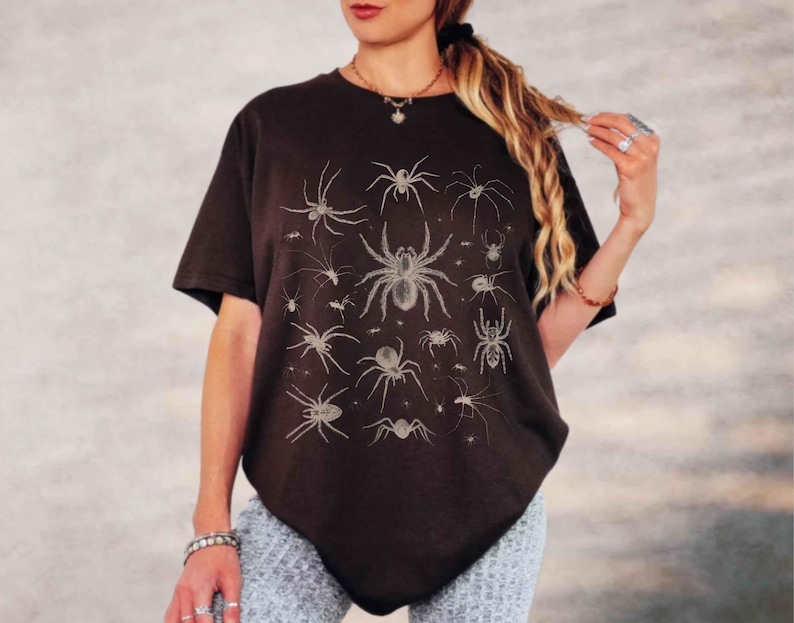May include: A black short-sleeved t-shirt featuring a graphic print of various spiders in a light beige colour. The design displays multiple spider species of different sizes arranged across the front of the garment, which has a crew neckline.