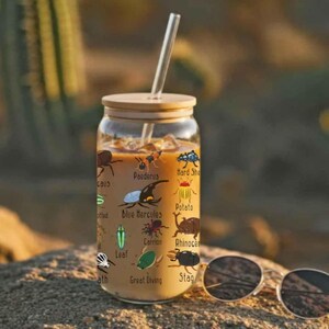 K&ouml;nnte beinhalten: Ein klares Glas mit Bambusdeckel und Glasstrohhalm, gef&uuml;llt mit einem hellbraunen Getr&auml;nk. Das Glas ist mit farbenfrohen Illustrationen verschiedener Insekten und ihren Namen verziert. Eine Sonnenbrille liegt auf der Oberfl&auml;che.