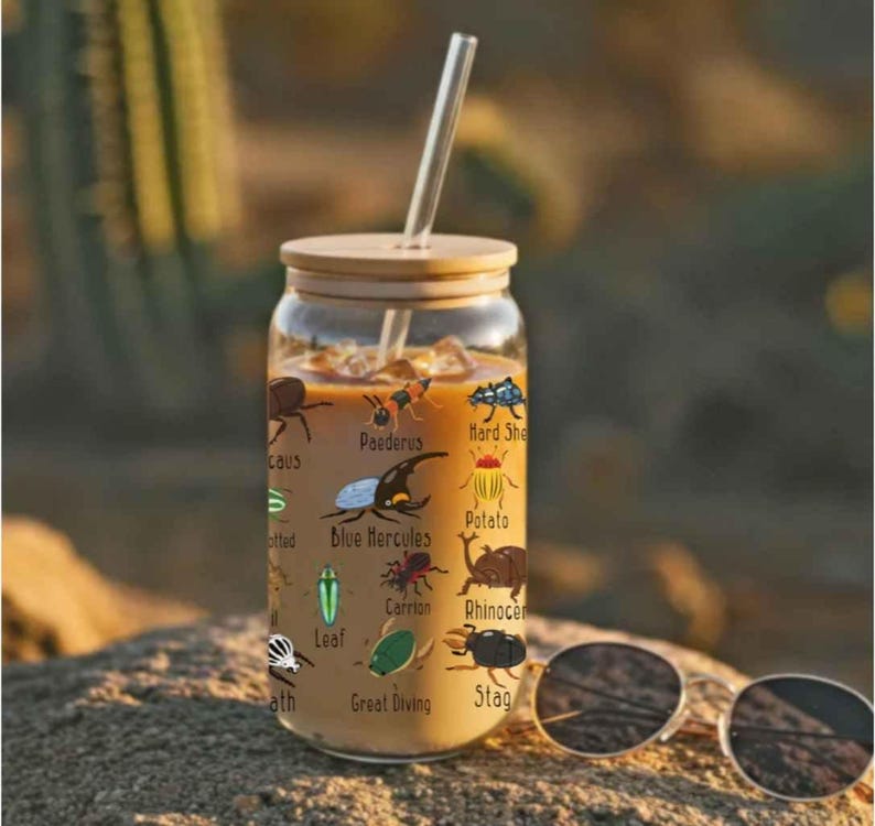 K&ouml;nnte beinhalten: Ein klares Glas mit Bambusdeckel und Glasstrohhalm, gef&uuml;llt mit einem hellbraunen Getr&auml;nk. Das Glas ist mit farbenfrohen Illustrationen verschiedener Insekten und ihren Namen verziert. Eine Sonnenbrille liegt auf der Oberfl&auml;che.