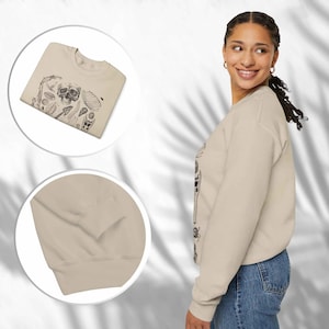 Op de afbeelding: Een beige sweatshirt met lange mouwen en een zwarte grafische print in inktstijl van een schedel, diverse schelpen en botanische elementen op de voorzijde. Het kledingstuk heeft een ronde hals en geribbelde boorden bij de mouwen.