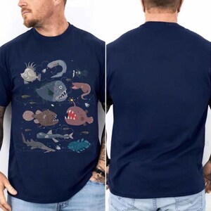 May include: A navy blue short-sleeve t-shirt featuring a graphic print of various deep-sea creatures, including an anglerfish, a sawfish, and other marine life. The back of the t-shirt is solid navy blue with no graphic print or additional design elements.