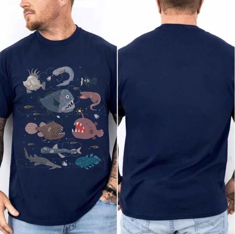 May include: A navy blue short-sleeve t-shirt featuring a graphic print of various deep-sea creatures, including an anglerfish, a sawfish, and other marine life. The back of the t-shirt is solid navy blue with no graphic print or additional design elements.