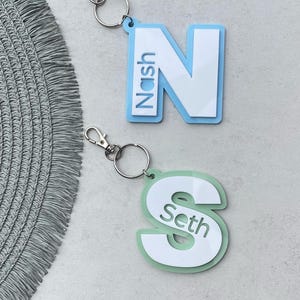 May include: Two personalised keychains featuring the letters N and S. The N is light blue with the name Nash written vertically on the left side. The S is light green with the name Seth written inside the letter.
