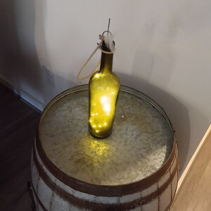 May include: A green glass bottle with a string handle and white lights inside, sitting on a rustic metal barrel table.