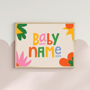 Puede incluir: Un estampado enmarcado con las palabras "BABY NAME" en letras coloridas y juguetonas. El dise&ntilde;o incluye formas florales y abstractas brillantes en naranja, rosa, verde y amarillo sobre un fondo crema. El marco es de color madera clara.