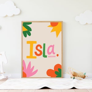 May include: A framed print with a beige background and colourful illustrations. The print features the name "Isla" in large, stylized letters with a yellow sun, green leaves, and pink and orange flowers.