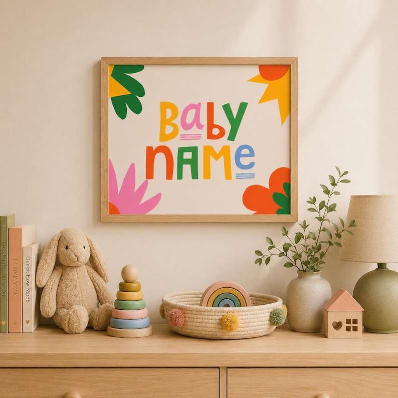 Puede incluir: Un cuadro enmarcado con las palabras "BABY NAME" en letras coloridas, rodeado de dise&ntilde;os florales y en forma de sol. Un conejito de peluche, un juguete apilable de madera y una cesta decorativa con un arco&iacute;ris est&aacute;n sobre una superficie de madera.