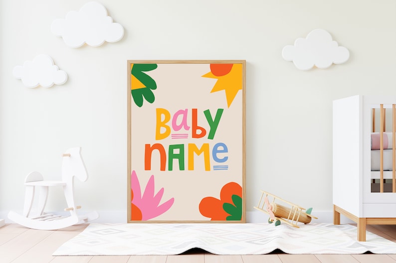 以下が含まれることがあります： カラフルで遊び心のある文字で「BABY NAME」と書かれた額入りのプリント。アートワークは、緑、オレンジ、ピンクの抽象的な花と太陽のデザインが特徴です。プリントは、白いベビーベッドとロッキングホースのある子供部屋に飾られています。