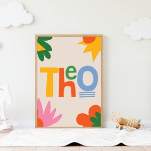 以下が含まれることがあります： カラフルで遊び心のある文字で「Theo」と書かれた額入りのアートプリント。オレンジ、黄色、緑、ピンクの抽象的な形が、ニュートラルな背景に配置されています。装飾的な雲のある子供部屋に飾られています。