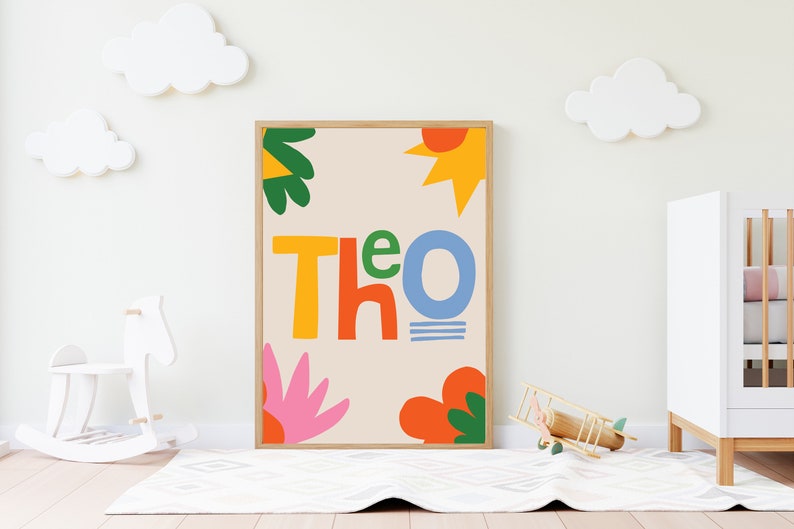 以下が含まれることがあります： カラフルで遊び心のある文字で「Theo」と書かれた額入りのアートプリント。オレンジ、黄色、緑、ピンクの抽象的な形が、ニュートラルな背景に配置されています。装飾的な雲のある子供部屋に飾られています。