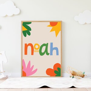 以下が含まれることがあります： カラフルで遊び心のある文字で「Noah」という名前が書かれた額入りのアートプリント。オレンジ、緑、黄色、ピンクの抽象的な花と太陽のデザインがベージュの背景に描かれています。プリントは子供部屋に飾られています。