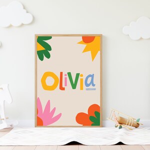 以下が含まれることがあります： カラフルで遊び心のある文字で「Olivia」という名前が書かれた額入りのアートプリント。オレンジ、ピンク、緑、黄色の抽象的な花や太陽のような形がベージュの背景に描かれています。子供部屋に飾られています。