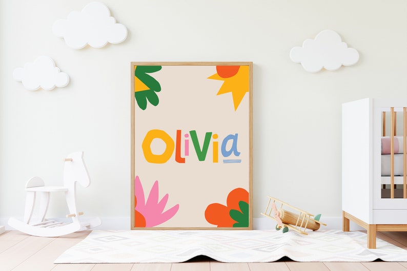以下が含まれることがあります： カラフルで遊び心のある文字で「Olivia」という名前が書かれた額入りのアートプリント。オレンジ、ピンク、緑、黄色の抽象的な花や太陽のような形がベージュの背景に描かれています。子供部屋に飾られています。