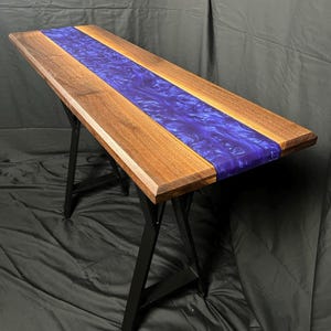 May include: A long, rectangular table with a walnut wood top and a blue resin inlay. The table is supported by black metal A-frame legs.