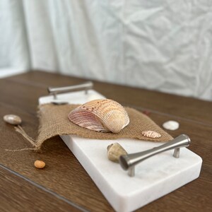 Könnte beinhalten: Weiße Marmor-Servierplatte mit zwei silbernen Griffen. Die Platte ist mit Muscheln und Jutegewebe verziert.
