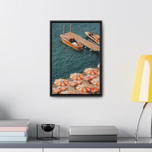 May include: A framed canvas print featuring a coastal scene. Two wooden boats are docked at a pier, with orange and white striped umbrellas and lounge chairs along the shore. The water is a deep blue.