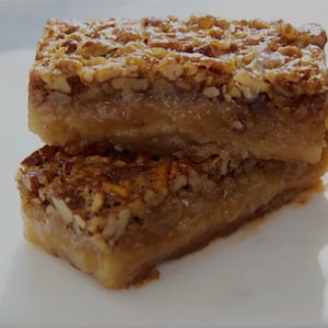 May include: Two pecan pie bars stacked on a white surface. The bars have a golden-brown base with a layer of caramel and a topping of chopped pecans. A dusting of powdered sugar adds a finishing touch. The image showcases a close-up of the dessert.