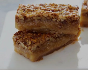 May include: Two pecan pie bars stacked on a white surface. The bars have a golden-brown base with a layer of caramel and a topping of chopped pecans. A dusting of powdered sugar adds a finishing touch. The image showcases a close-up of the dessert.