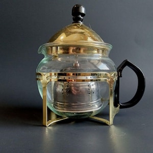 May include: A clear glass teapot featuring a gold-tone metal frame and base, a black handle, and a black knob on the lid. Inside the glass body sits a removable stainless steel tea infuser with small circular perforations for steeping loose tea leaves.
