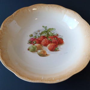 May include: A round, off-white ceramic serving bowl with a scalloped edge. The bowl features a painted illustration of ripe strawberries and gooseberries. The rim has a light brown color. A decorative piece for serving fruit.