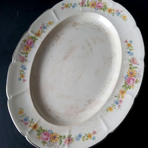 May include: Oval serving platter with a scalloped edge and floral design. The off-white platter features a border of pink, yellow, and blue flowers. The rim has a silver trim.