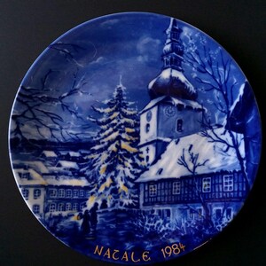 May include: A blue and white decorative plate featuring a winter village scene with a church, snow-covered buildings, and a Christmas tree. The plate has the word "NATALE" and the year "1984" in gold lettering.