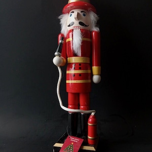 May include: A red wooden nutcracker soldier with a tall hat and white hair and mustache. The nutcracker has a black and gold base and holds a black and white object. A red tag with a Christmas tree and the words "Holiday Magic" is attached.