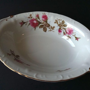 May include: Oval-shaped, off-white serving dish with a gold rim and handles. The dish is decorated with pink roses and brown leaves. The edges have a scalloped design.