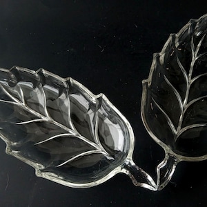 May include: Two clear glass leaf-shaped serving dishes. The dishes have detailed vein patterns and a glossy finish. The leaves are connected at the base, creating a unique serving piece. The dishes are ideal for serving snacks or small portions.