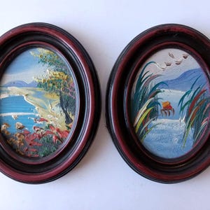 May include: Two oval framed oil paintings. One depicts a coastal scene with a blue ocean, sandy shore, and green foliage. The other shows a river scene with tall grass, a small boat, and birds in the sky. The frames are dark red.