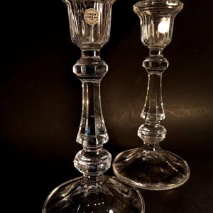 May include: Two clear crystal candle holders. The candle holders have a round base, a faceted stem, and a flared top. One candle holder has a label that reads "Cristal d'Arques France". The background is black.