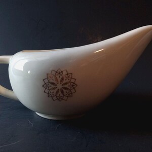 May include: White ceramic gravy boat with a handle and a spout. A brown floral design decorates the side of the gravy boat. The gravy boat is on a dark surface.
