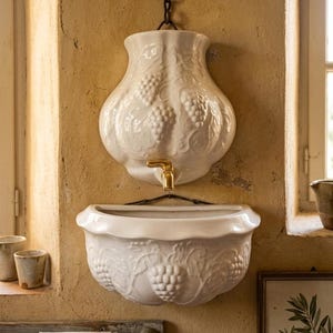 May include: White ceramic wall-mounted sink with a decorative grape vine design. The sink has a brass faucet and is mounted above a matching basin. The sink is hanging from a chain and is set against a textured, yellow wall.